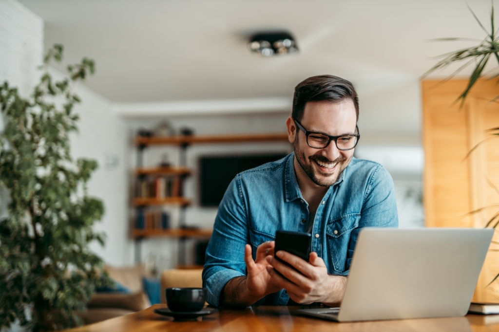 Smiling Man Sitting at Desk With Phone, Laptop, and Coffee