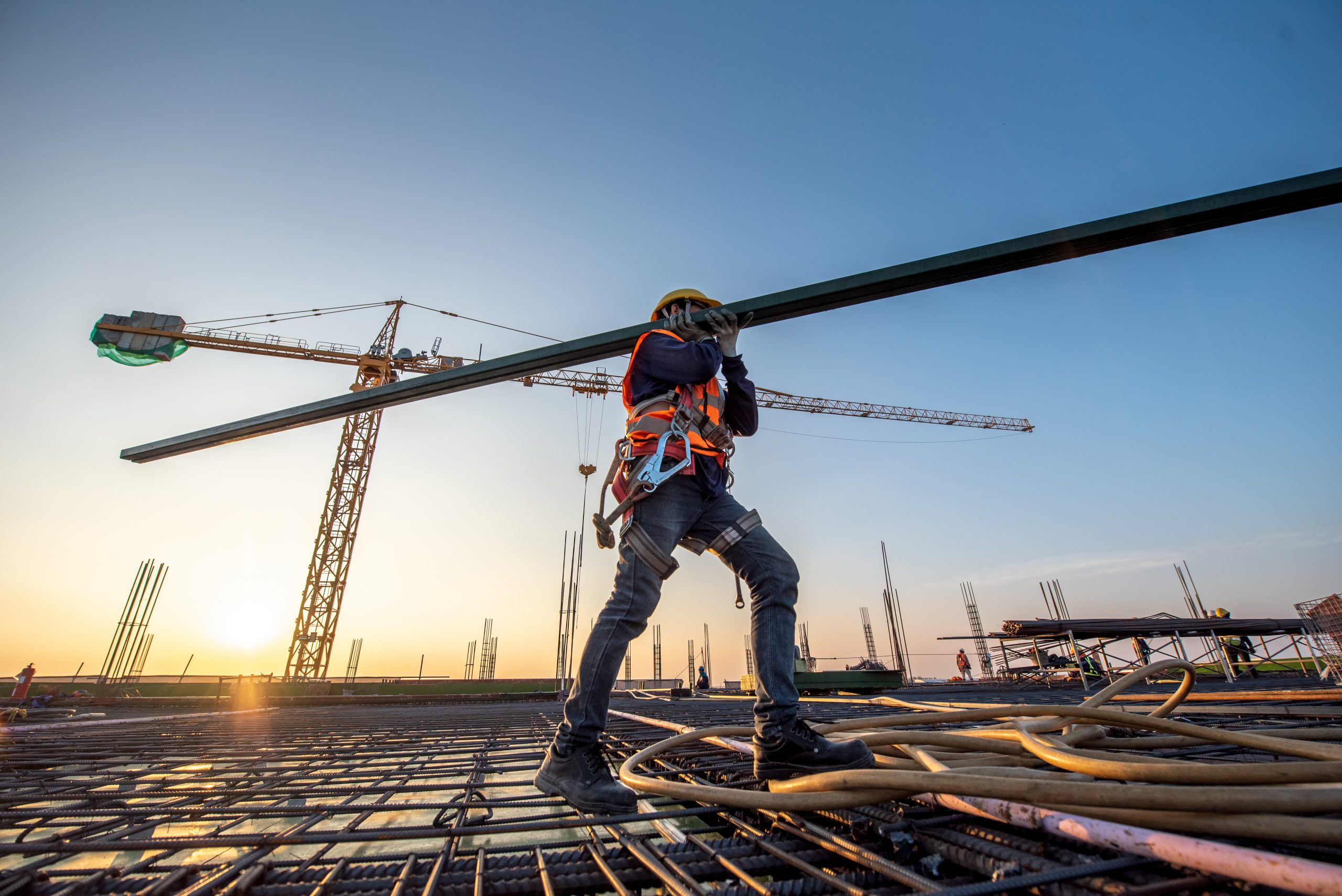 A Construction Worker Carrying Metal Beam With Crane in Background
