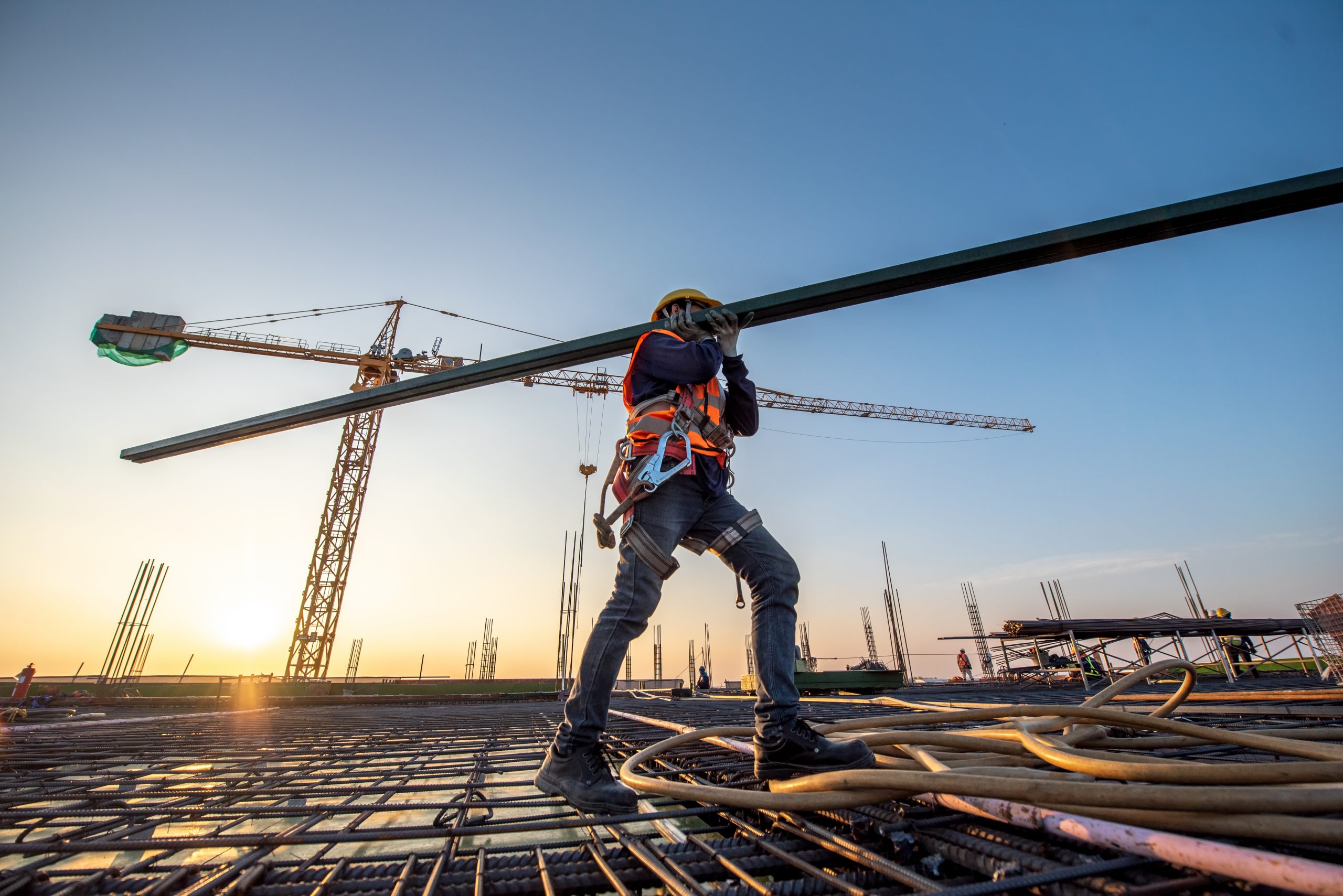 A Construction Worker Carrying Metal Beam With Crane in Background A Construction Worker Carrying Metal Beam With Crane in Background