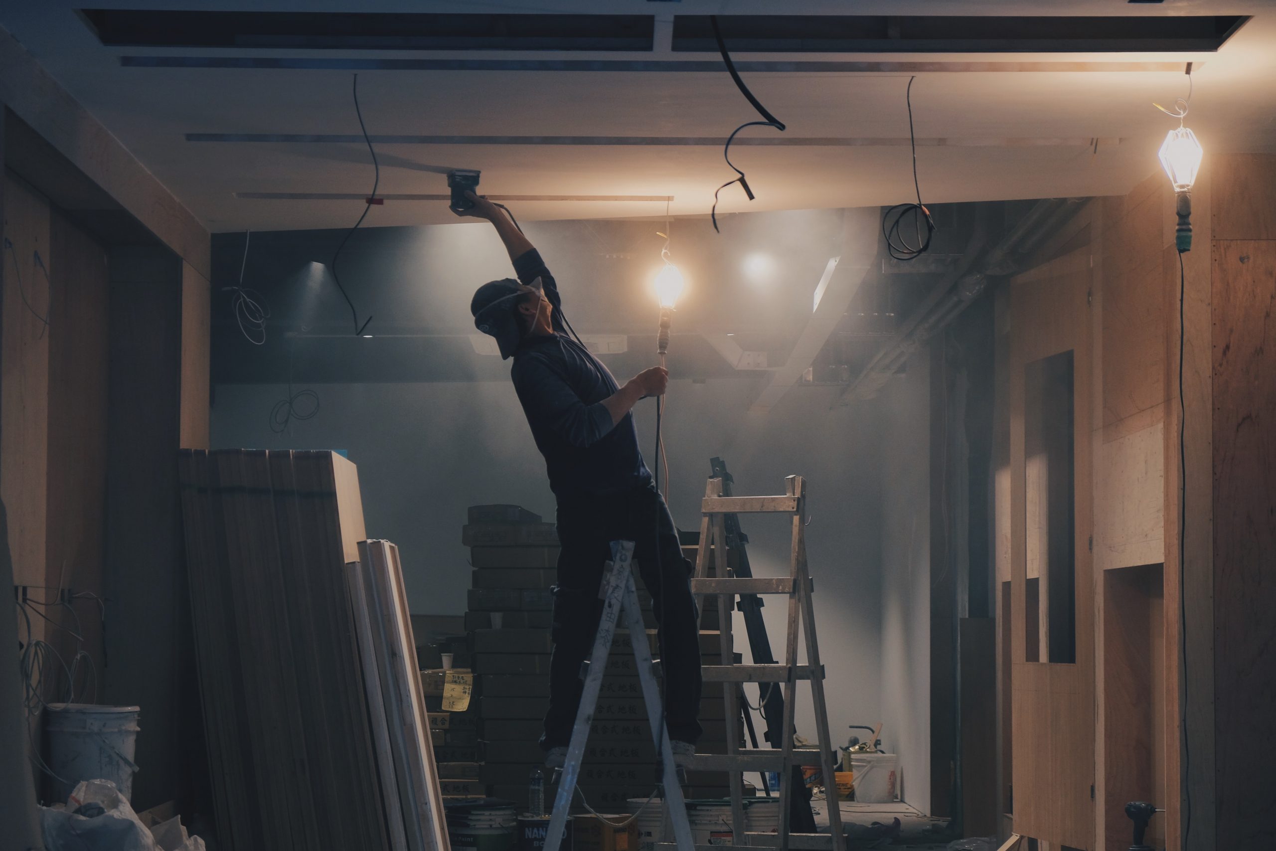 Man Straddling Ladder While Sanding Ceiling In Dark Dusty Room
