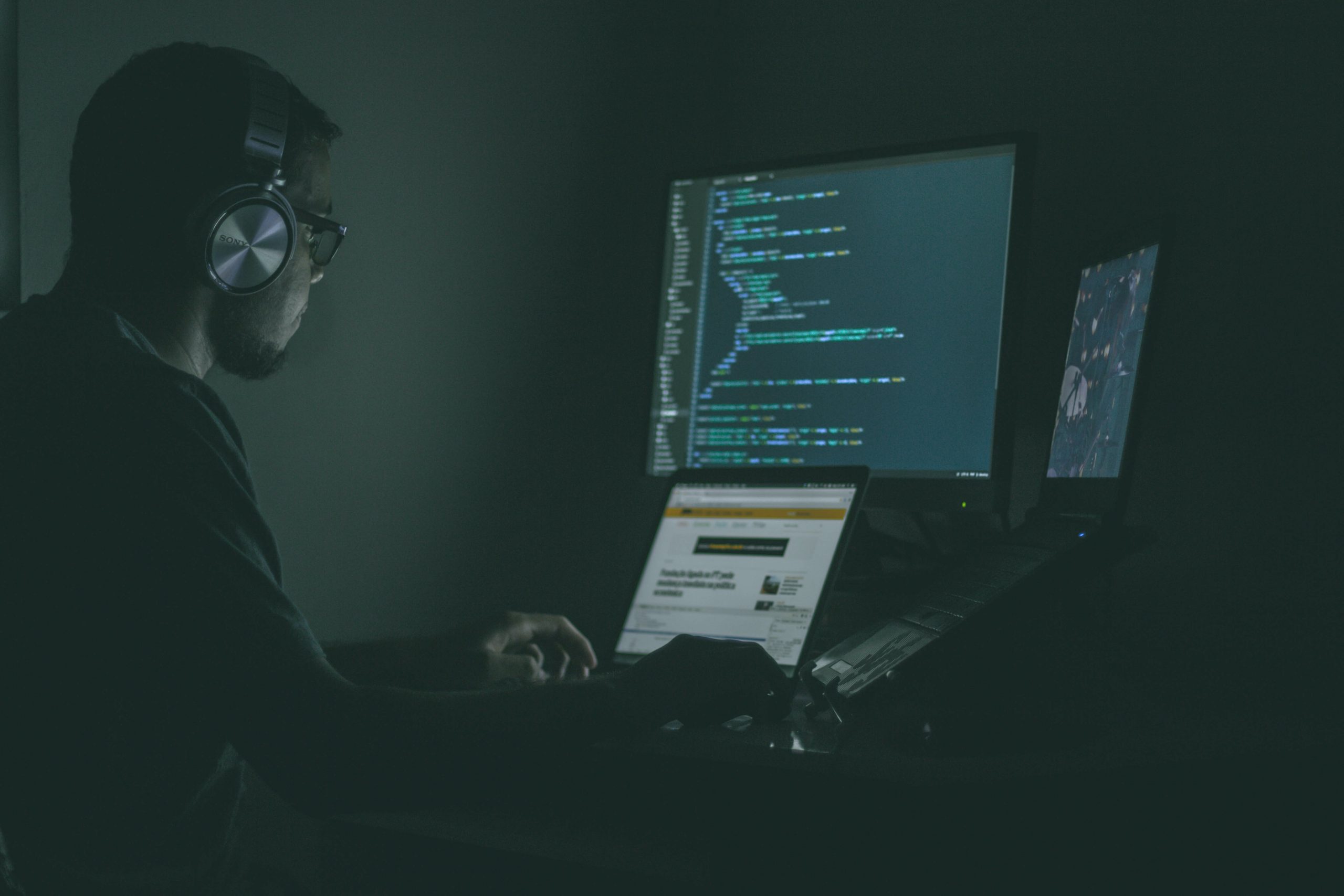 Man Wearing Headphones Coding on Three Computer Screens In Dark Room Man Wearing Headphones Coding on Three Computer Screens In Dark Room