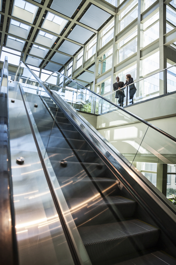 Business man and woman talking at the top of an escalator in a large glassed in open lobby area..,USA Tower Street Insurance Loss Control OSHA Safe and Sound Week